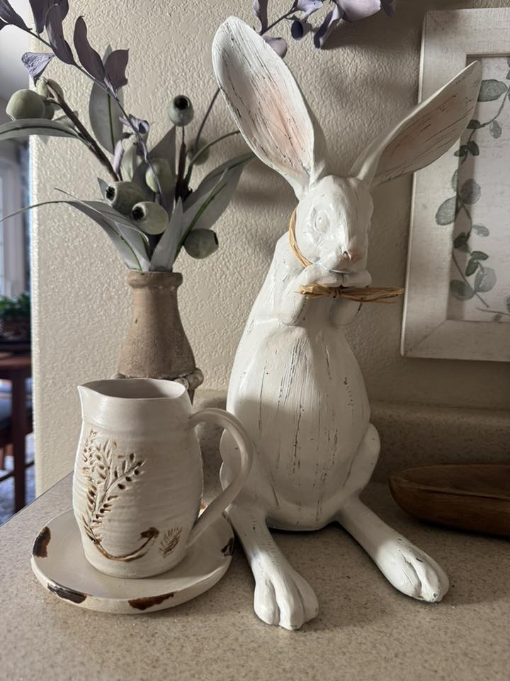 The mug, handthrown by The Potter's Wife Pottery company, features a vibrant red glaze with a textured, speckled finish. A prominent, raised heart design in a natural beige tone adorns the front, crafted with a twisted, rope-like border that adds a rustic touch. The mug has a sturdy handle and sits on a defined base, characteristic of hand thrown pottery. The overall design is warm and artisanal, blending bold color with delicate detailing.