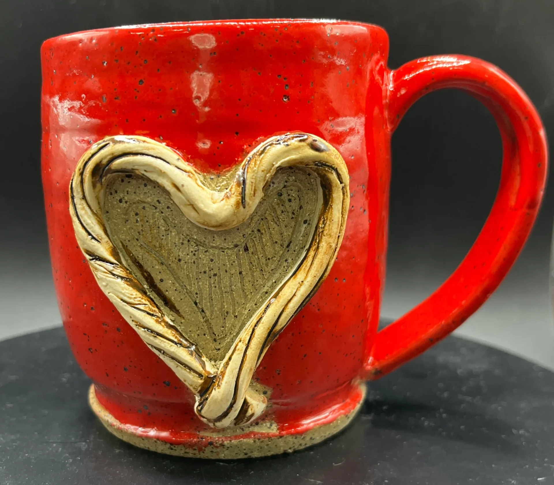 The mug, handthrown by The Potter's Wife Pottery company, features a vibrant red glaze with a textured, speckled finish. A prominent, raised heart design in a natural beige tone adorns the front, crafted with a twisted, rope-like border that adds a rustic touch. The mug has a sturdy handle and sits on a defined base, characteristic of hand thrown pottery. The overall design is warm and artisanal, blending bold color with delicate detailing.